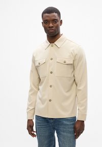 Μπεζ πουκάμισο με κουμπιά, διαθέτει δύο τσέπες στο στήθος, με λοξό γιακά και μακριά μανίκια, φτιαγμένο από απαλό ύφασμα με δομημένη εφαρμογή.