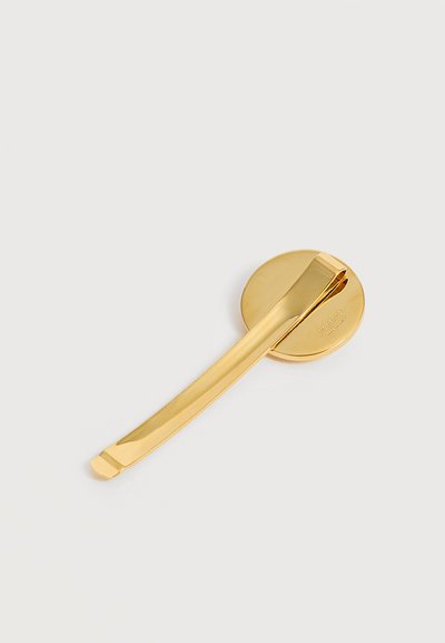 Barrette à cheveux en métal de couleur dorée avec une base ronde et un long fermoir courbé, placée sur un fond gris clair.