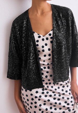 Femme porte une veste noire à sequins sur une robe blanche à gros pois noirs, debout devant un fond clair et uni.