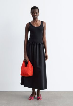 Femme portant une robe noire sans manches, des colliers et boucles d'oreilles en or, tenant un sac à main rouge, et portant des talons ouverts roses, debout sur un sol en béton.