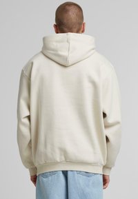 Krämfärgad huvtröja med en rymlig huva, ribbade cuffs och fåll. Har en slät textur och avslappnad passform, tillsammans med ljusblå jeans.