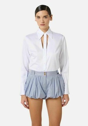 Donna che indossa una camicia bianca a maniche lunghe con bottoni e una spaccatura frontale, abbinata a pantaloncini a righe blu a vita alta con effetto gonfio, in piedi davanti a uno sfondo semplice.