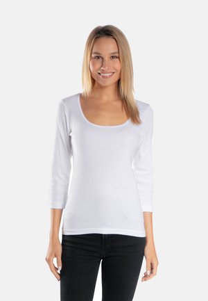 Uśmiechnięta kobieta o prostych blond włosach ma na sobie białą, długorękawową bluzkę z dekoltem w kształcie litery U oraz czarne spodnie, stojąc na jednolitym białym tle.