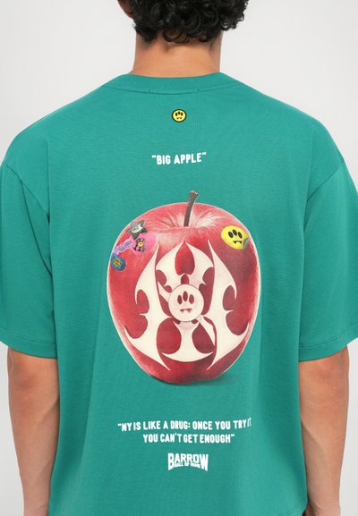 Green cotton t-shirt featuring a large red apple graphic with a white design and various colorful stickers, text above and below the apple.