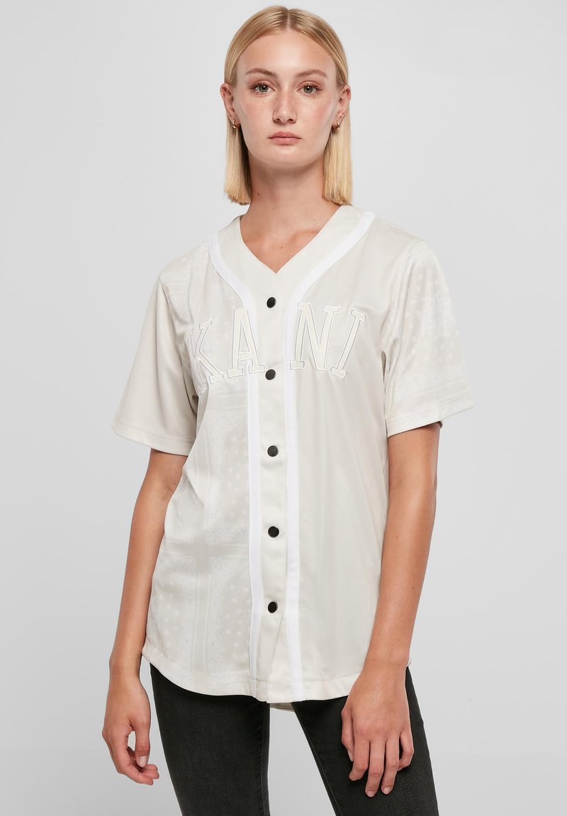 Kortærmet, button-up skjorte i lys beige, med V-hals, hvide detaljer og et grafisk "KANI" på forsiden, samt en glat tekstur.