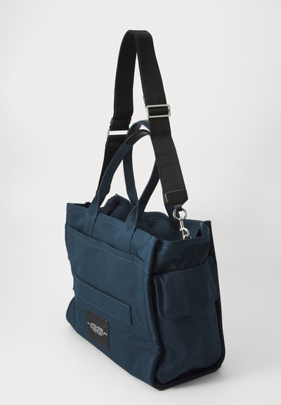 Mala de lona azul marinho com duas alças curtas e uma alça preta destacável. Apresenta bolsos frontal e lateral, e um rótulo preto na frente.