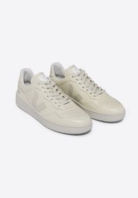 Beiga lädersneakers med perforerad tå, platta snörningar och en vit logotyp på tungan. Platt gummisula och diskret varumärkesinplacering på sidan.