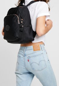 Frau trägt hellblaue Levi's Jeans und ein weißes, kurzes Shirt, während sie einen schwarzen Rucksack mit roségoldenen Reißverschlüssen über einer Schulter trägt.