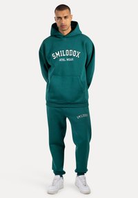 Felpa cappuccio color teal con testo bianco "SMILODOX ATHL WEAR", tasca a marsupio, abbinata a pantaloni coordinati. Entrambi gli articoli presentano un tessuto morbido e una vestibilità rilassata.