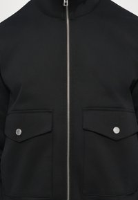 Jachetă neagră cu guler înalt, dotată cu un fermoar argintiu și două buzunare frontale cu clapetă și capse. Material neted cu un design structurat.