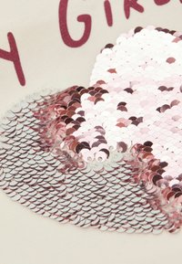 Paillettenaufnäher mit einem Farbübergang aus silbernen und rosaroten kreisförmigen Pailletten auf einem hellbeigen Stoffhintergrund mit Text darüber.