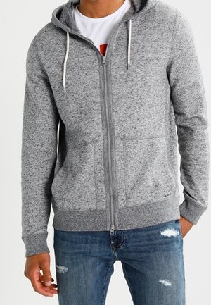 Mann trägt ein graues Kapuzensweatshirt mit Reißverschluss und abgenutzte blaue Jeans, steht mit der linken Hand in der Tasche.