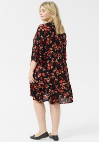 Vestido negro con flores rojas, de mangas cortas, falda de volantes y tela ligera. Llevado con zapatos planos negros, visto desde la parte posterior.