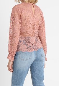 Lyserød blondebluse med lange ærmer, der har en bølget kant og design med nøglehul bagpå, parret med lyseblå denimjeans.