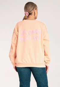 Jasnoróżowy, oversize'owy sweatshirt z napisem "SECONDO ME CI STA" w teksturowej purpurowej haftowanej czcionce na plecach. Miękka tkanina, ściągacze przy mankietach i u dołu.