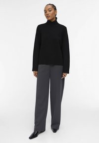 Maglione nero a maniche lunghe con collo alto, abbinato a pantaloni grigi scuri larghi e scarpe nere lucide, su uno sfondo neutro.