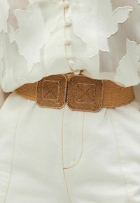 Gewebter, tanfarbener Gürtel mit quadratischen, geflochtenen Schnallen-Details; getragen über einer weißen Bluse mit floralem Muster; weiche Textur und neutrale Farben.