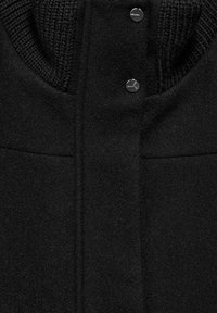 Svart ullblandad kappa med ribbad krage, slät textur och blanka metallknappar för stängning. Minimalistisk design med två sidofickor.