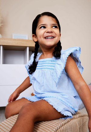 Smilende ung pige med fletninger iført en blå stribet kjole sidder på en beige tekstureret skammel i et minimalistisk rum.