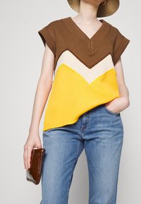 Hnědý, krémový a žlutý pletený top má chevronový vzor, hluboký V-výstřih a krátké rukávy. Modelka drží hnědou kabelku.