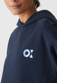 Felpa blu navy con una texture morbida, cappuccio con cordino e un logo ricamato blu chiaro "OX" sul petto, che presenta un design geometrico.