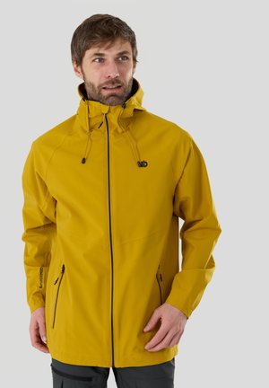 Homme portant une veste imperméable jaune moutarde avec fermeture éclair frontale et pantalon noir, regardant légèrement vers la gauche sur un fond gris.