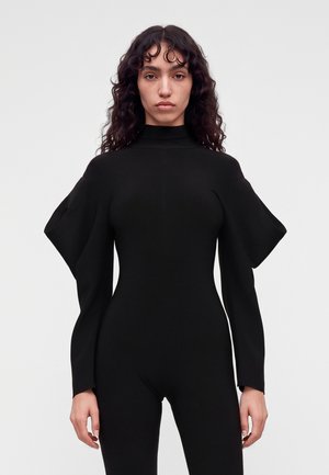 Femme aux cheveux bouclés et foncés portant une combinaison noire ajustée avec un col montant et de larges manches structurées aux épaules sur fond blanc.