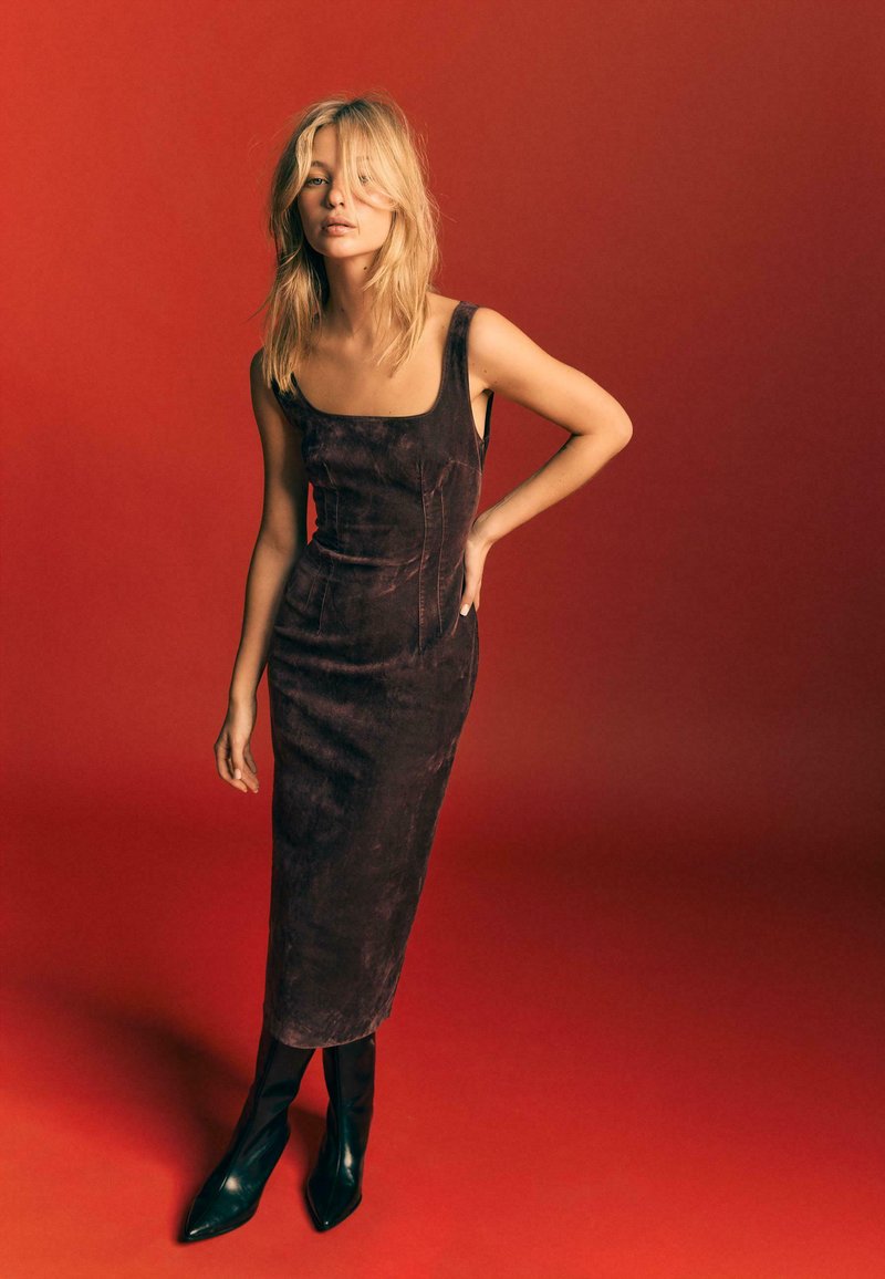 Robe en daim marron avec un décolleté carré et une silhouette ajustée, associée à des bottes noires à bouts pointus, sur un fond rouge uni.