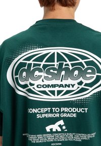 Zielona bawełniana koszulka z dużym białym logo z motywem globu oraz tekstem, w tym "DC Shoe Company" i "Superior Grade."