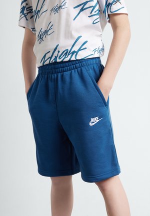 Barn iført blå Nike shorts og en hvid t-shirt med blå "Flight" tekst, hænderne i lommerne, mod en ensartet lys baggrund.