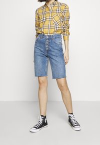 Camicia a quadri gialla con bottoni e maniche lunghe, abbinata a pantaloni corti di denim blu con chiusura a bottoni e orlo sfilacciato, indossata con scarpe da ginnastica nere in tela.