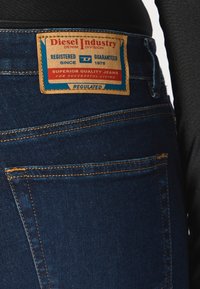 Σκούρο μπλε τζιν παντελόνι με εμφανή καφέ δερμάτινη ετικέτα που περιλαμβάνει κόκκινη και μπλε σήμανση, ανθεκτικά ραψίματα και κλασικό σχέδιο με πέντε τσέπες.