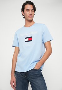 Νεαρός άνδρας στέκεται με το ένα χέρι στην τσέπη, φορώντας ανοιχτό μπλε T-shirt Tommy Hilfiger και σκούρα μπλε τζιν, μπροστά σε λευκό φόντο.