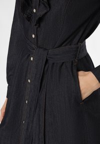 Denimkleid in Dunkelblau mit Rüschensaum, silbernen Knöpfen, tailliertem Gürtel und kontrastierenden orangefarbenen Nähten.