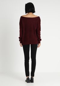 Pull tricot bordeaux à épaules dénudées avec des longues manches, texture côtelée et coupe décontractée, associé à un pantalon noir slim et des chaussures à talons carrés noires.
