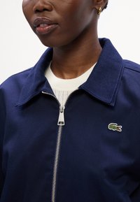 Mornarsko modra zip-up jakna s strukturiranim ovratnikom, ki ima majhen zeleni vezen logotip krokodila in srebrno zadrgo.