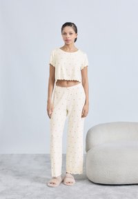 Σετ πιτζάμας ανοιχτού κίτρινου χρώματος με κοντό τοπ και φαρδιά παντελόνια, και τα δύο με μικρά κόκκινα μοτίβα καρδιάς. Μαλακό ύφασμα και στρογγυλεμένες άκρες.