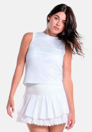 Vrouw met lang donker haar die een mouwloos wit topje en een gelaagde witte rok met gegolfde randen draagt tegen een effen achtergrond.