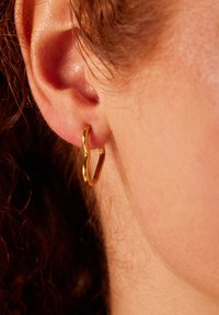 Gouden gedraaide creool met een gepolijste afwerking en een moderne, gebogen vorm, bevestigd aan een oor. Close-up van de details van de oorbel.