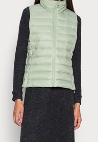 Gilet imbottito verde chiaro con colletto alto, chiusura a zip e motivo trapuntato orizzontale. Indossato sopra un vestito maglione scuro.