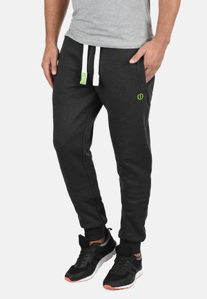 Dunkelgraue Jogginghose aus weichem Stoff, mit weißen Kordeln, seitlichen Taschen und einem kleinen grünen Logo auf der Vorderseite. Schwarze Sportschuhe.