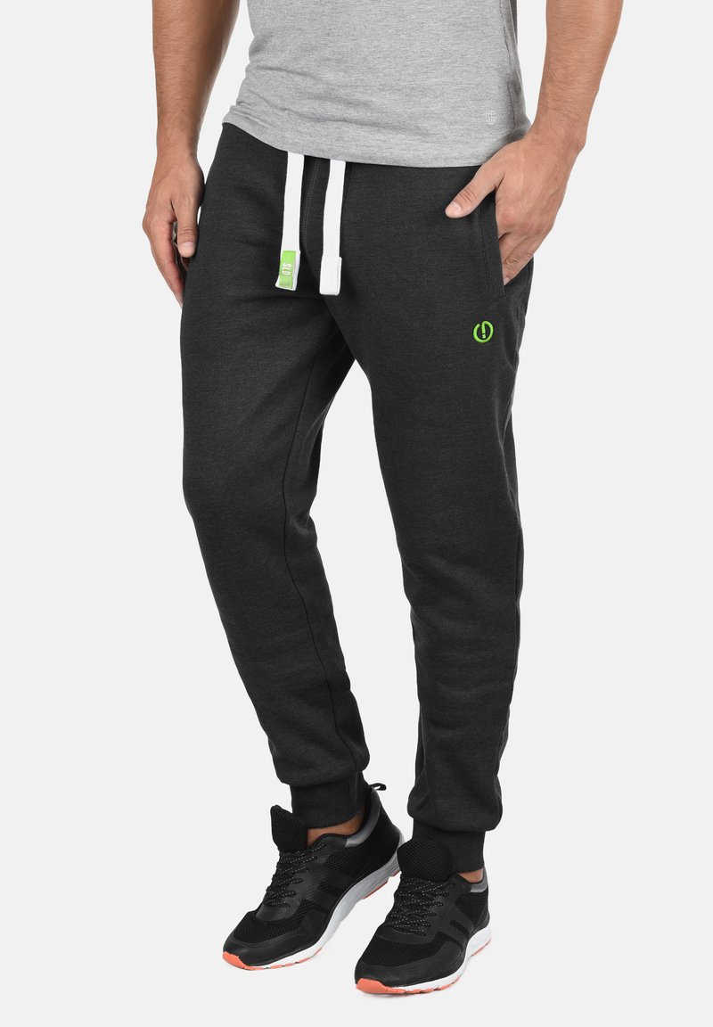 Dark gray joggers made of soft fabric, featuring white drawstrings, side pockets, and a small green logo on the front. Black athletic shoes.