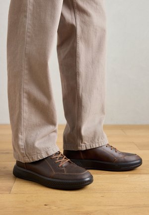 Bruine leren schoenen met een gestructureerd oppervlak en lichtbruine veters. Ronde neus en een laag profiel zwarte zool. Gepaarde met beige broek.