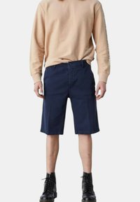 Pantaloni corti blu navy realizzati in tessuto misto cotone, caratterizzati da un design frontale piatto, cinque tasche e dettagli cuciti, abbinati a una felpa beige.