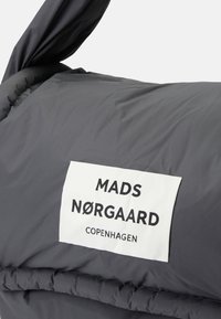 Sötét szürke steppelt kabát fehér téglalap alakú címkével, amelyen vastag fekete betűkkel szerepel a "MADS NØRGAARD COPENHAGEN" felirat.