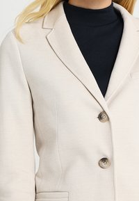 Beige blazer na míru vyrobený z texturované tkaniny, s klopovým límcem a dvěma velkými knoflíky. Nositý přes černý rolák.