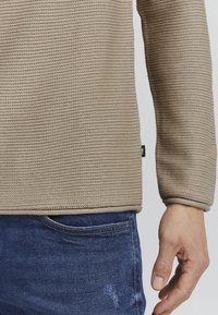 Beige gestrickter Pullover mit einem strukturierten Muster und gerippten Bündchen. Über blauen Jeans getragen, die einen ordentlich abgeschlossenen Saum und ein seitliches Etikett zeigen.