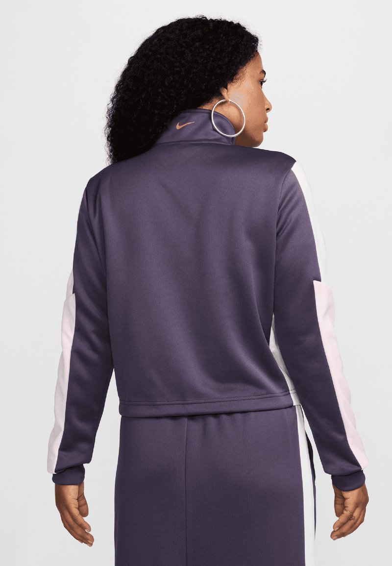 Chaqueta deportiva morada con paneles laterales blancos y rosas, hecha de una tela suave. Presenta un cuello alto y un detalle de logo cerca del cuello.