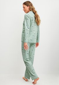 Woman in green pajama set with polar bear and snowflake pattern, standing barefoot against a white background, facing sideways.