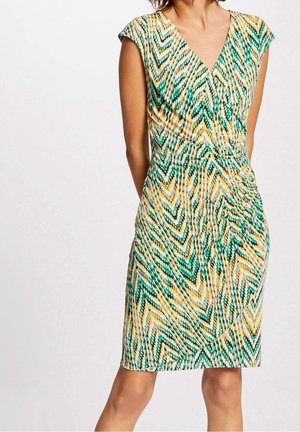 Mujer con un vestido ajustado hasta la rodilla, con escote en V y patrón de zigzag multicolor en tonos verdes, amarillos y negros.